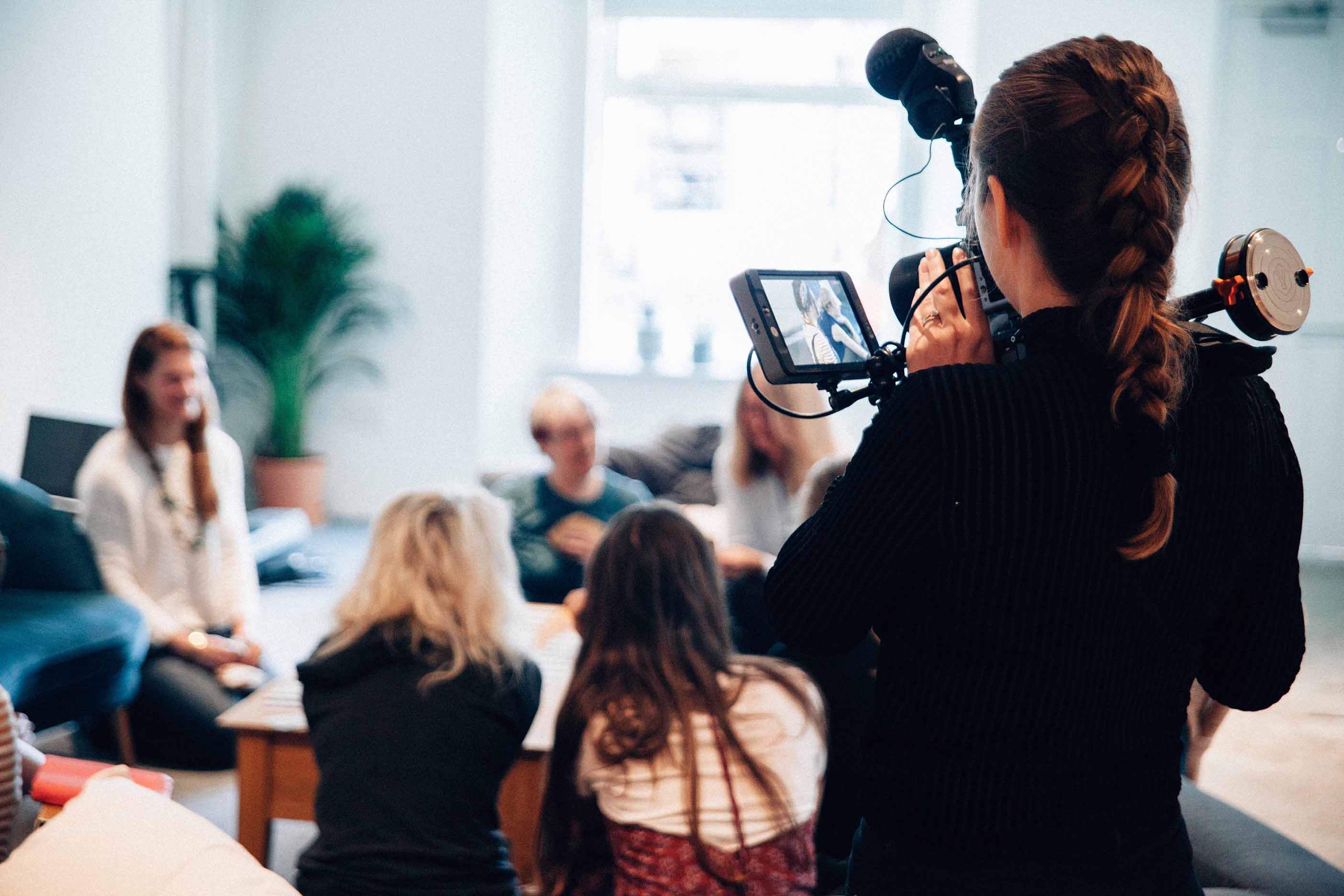 Woman holding a camera filming an interview. Interview subject and interviewer are out of focus.