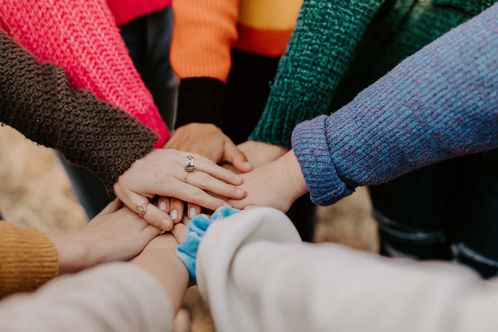 People putting their hands together to signify teamwork and collaboration. Many of them are wearing colourful knitwear.