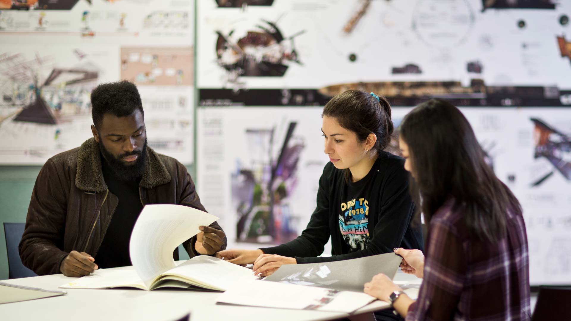 Three students sit around a table looking at pictures and books. In the background the wall is full of blurred out drawings.